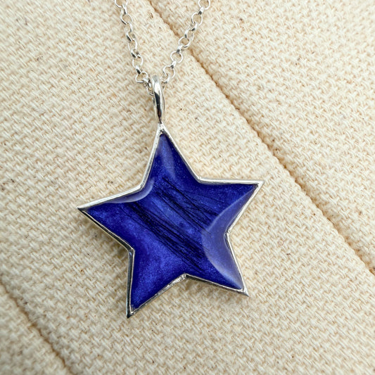 A close-up of a delicate sterling silver chain necklace with a five-pointed star pendant featuring a deep blue inlay and hair inclusion.