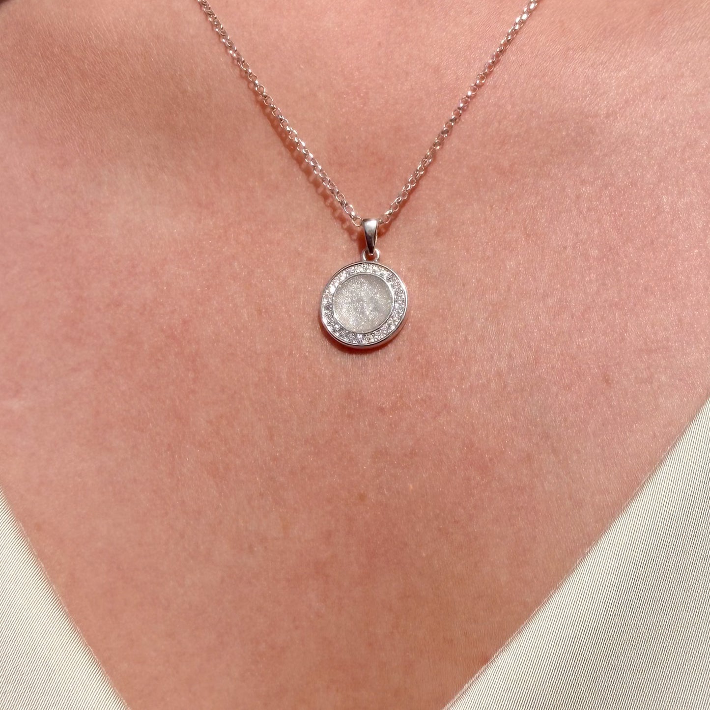 A close-up of a person's neck wearing a silver-toned necklace with a round pendant. The pendant features a textured, rough stone surrounded by a halo of small clear stones, suspended from a delicate chain.
