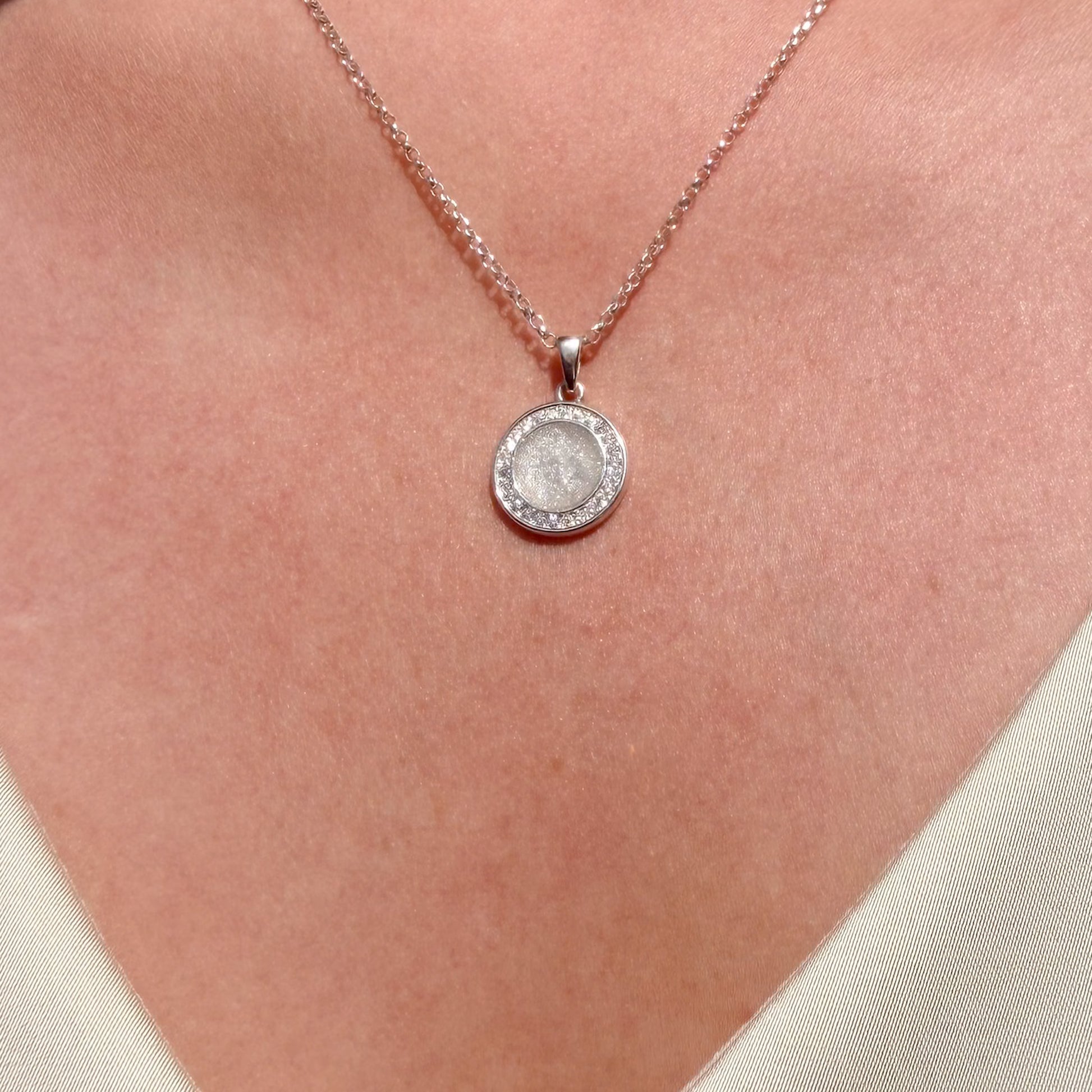 A close-up of a person's neck wearing a silver-toned necklace with a round pendant. The pendant features a textured, rough stone surrounded by a halo of small clear stones, suspended from a delicate chain.