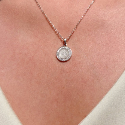 A close-up of a person's neck wearing a silver-toned necklace with a round pendant. The pendant features a textured, rough stone surrounded by a halo of small clear stones, suspended from a delicate chain.