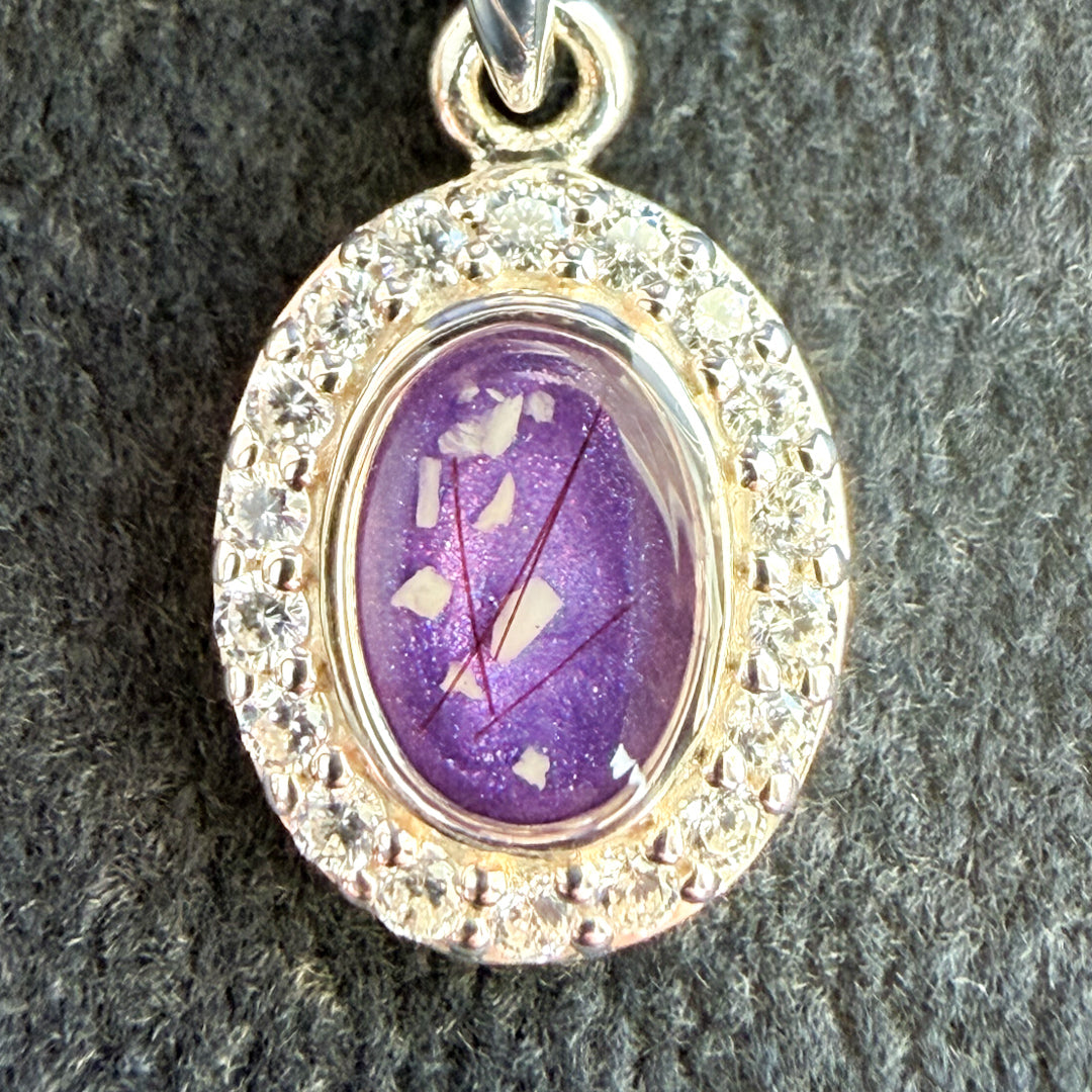 A close up of a sterling silver oval pendant surrounded by cubic zirconia stones on a sterling silver necklace featuring a purple amethyst center stone with visible ashes and hair strands. 