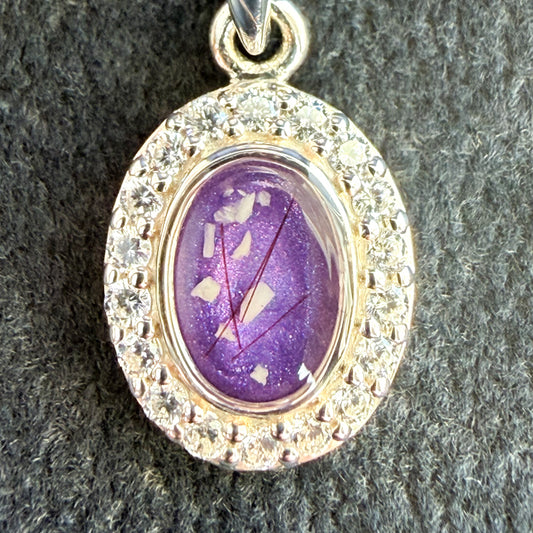 A close up of a sterling silver oval pendant surrounded by cubic zirconia stones on a sterling silver necklace featuring a purple amethyst center stone with visible ashes and hair strands. 