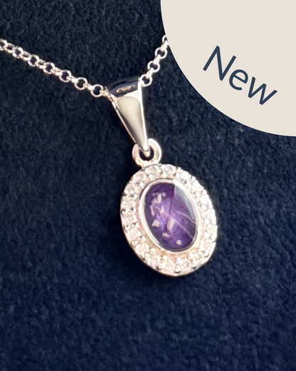 A sterling silver oval pendant surrounded by cubic zirconia stones on a sterling silver necklace featuring a purple amethyst center stone with visible ashes and hair strands. 
