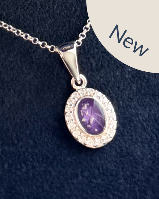 A sterling silver oval pendant surrounded by cubic zirconia stones on a sterling silver necklace featuring a purple amethyst center stone with visible ashes and hair strands. 