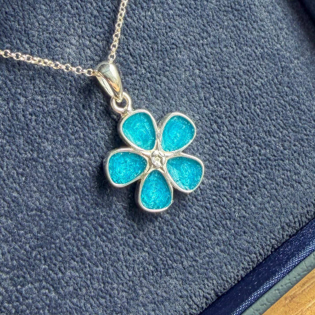 A silver flower-shaped pendant necklace with five bright turquoise blue enamel petals and a small clear center stone, resting on a textured dark blue surface.