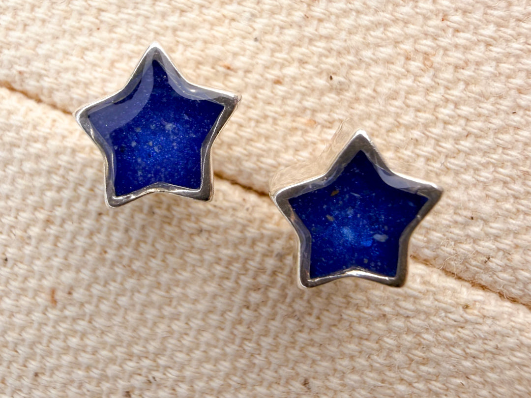 A pair of sterling silver star-shaped stud earrings with deep blue inlay and visible ash, displayed on a textured beige fabric background.