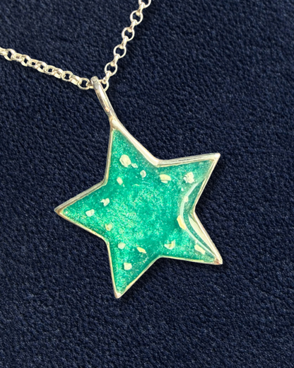 A five-pointed star pendant necklace with a textured green enamel center and small gold speckles, hanging from a silver-toned chain against a dark navy blue textured background.