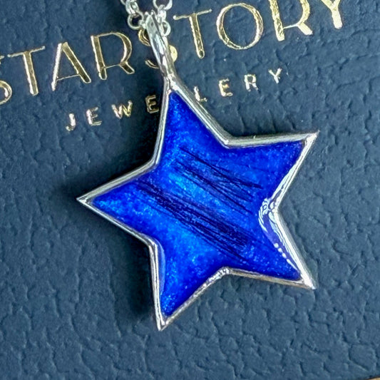 A five-pointed star pendant necklace with a vibrant blue inlay and silver metal border, resting on a textured dark blue surface with gold 'STARSTORY JEWELLERY' branding visible.