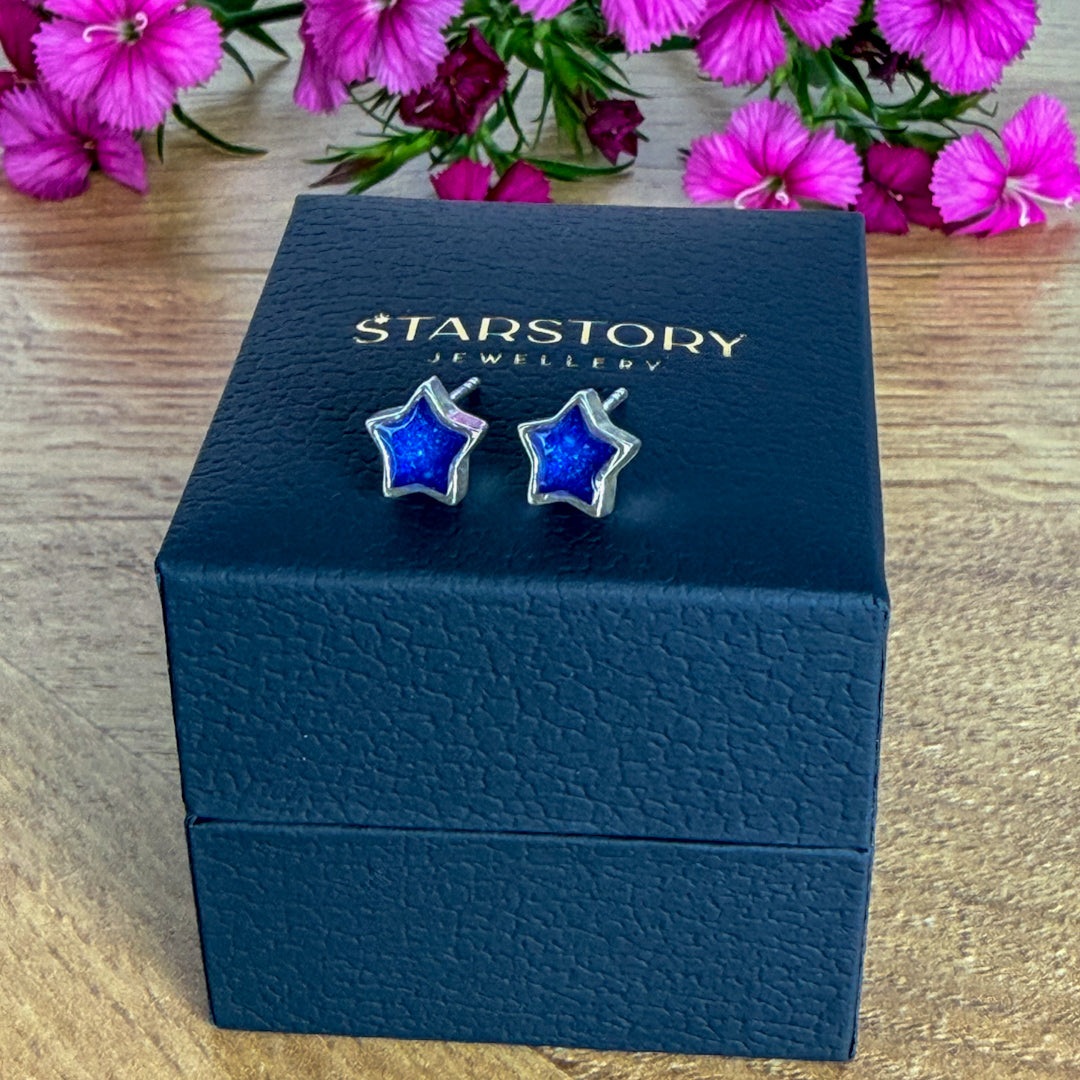 Star-shaped stud earrings with blue centers and silver borders, with visible blended ashes within. They are displayed on a dark blue Starstory Jewellery box on a wooden surface with pink flowers in the background.