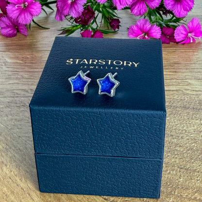 Star-shaped stud earrings with blue centers and silver borders, with visible blended ashes within. They are displayed on a dark blue Starstory Jewellery box on a wooden surface with pink flowers in the background.