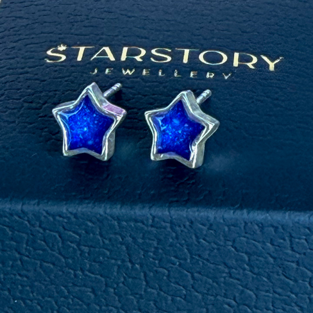 Star-shaped stud earrings with blue centers and silver borders, with visible blended ashes within. They are displayed on a dark blue Starstory Jewellery box on a wooden surface with pink flowers in the background.