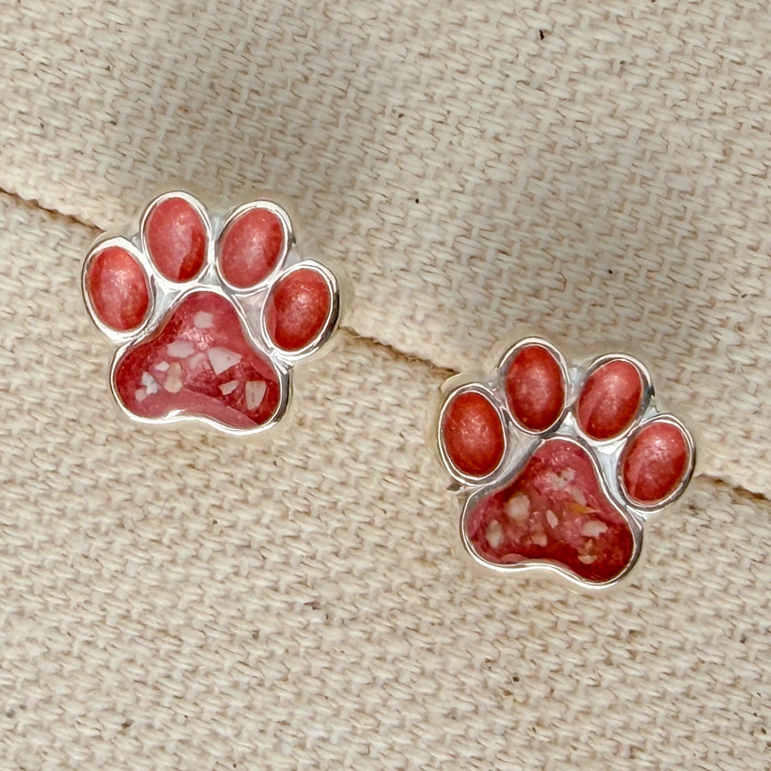 A pair of sterling silver paw print stud earrings filled with blush pink resin and small flecks ash, displayed on a textured beige woven fabric background.
