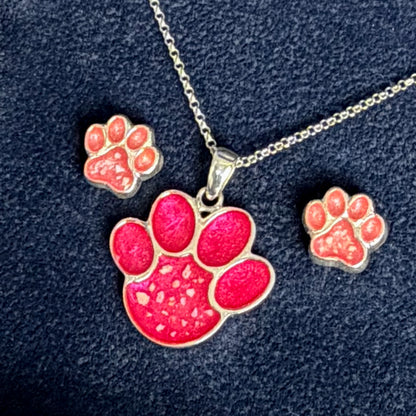 A silver-tone paw print pendant necklace with a matching stud earring and small rectangular charm, all featuring bright pink and coral-red paw prints on a dark navy textured background.