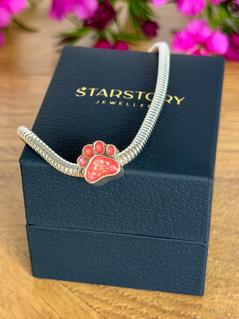 A silver-toned snake chain bracelet with a pink paw print charm with visible ashes, displayed on a dark blue Starstory Jewellery box on a wooden surface with blurred pink flowers in the background.