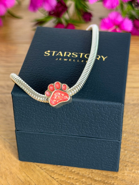 A silver-toned snake chain bracelet with a pink paw print charm with visible ashes, displayed on a dark blue Starstory Jewellery box on a wooden surface with blurred pink flowers in the background.