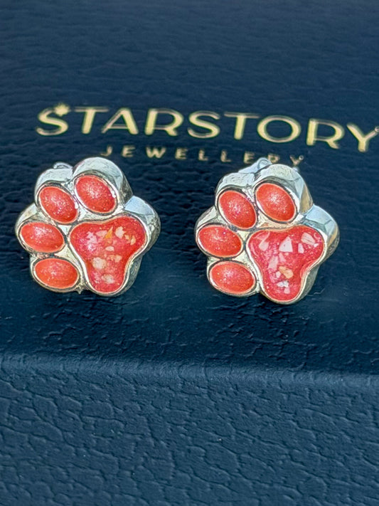 A pair of silver-toned stud earrings shaped like pink paw prints, displayed on top of a dark blue Starstory Jewellery box on a wooden surface with blurred pink flowers in the background.