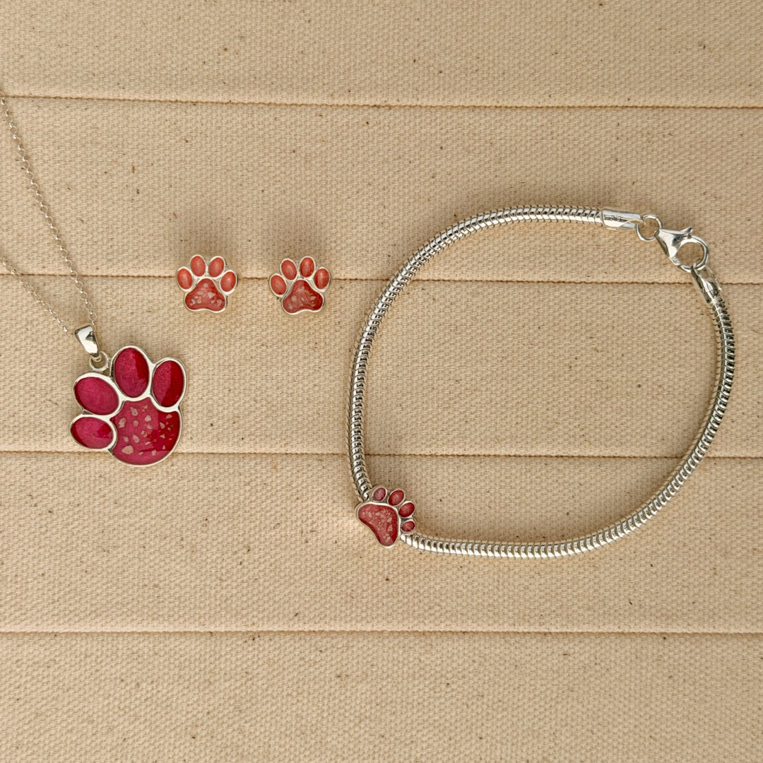 A sterling silver snake chain bracelet with charm, necklace and earrings featuring a paw print filled with textured pink resin and visible inclusions. 
