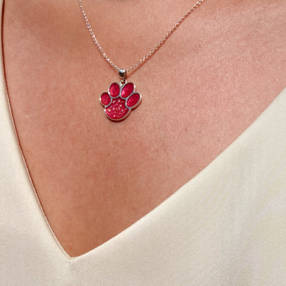 A close-up of a lady wearing a delicate sterling silver chain necklace with a rosey pink paw print pendant with visible ash, styled with a cream V-neck top.
