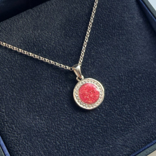 A silver halo pendant necklace featuring a round pink center with blended ashes surrounded by a halo of small clear stones, displayed on a textured dark blue background.