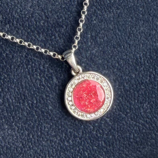 A silver halo pendant necklace featuring a round pink center with blended ashes surrounded by a halo of small clear stones, displayed on a textured dark blue background.