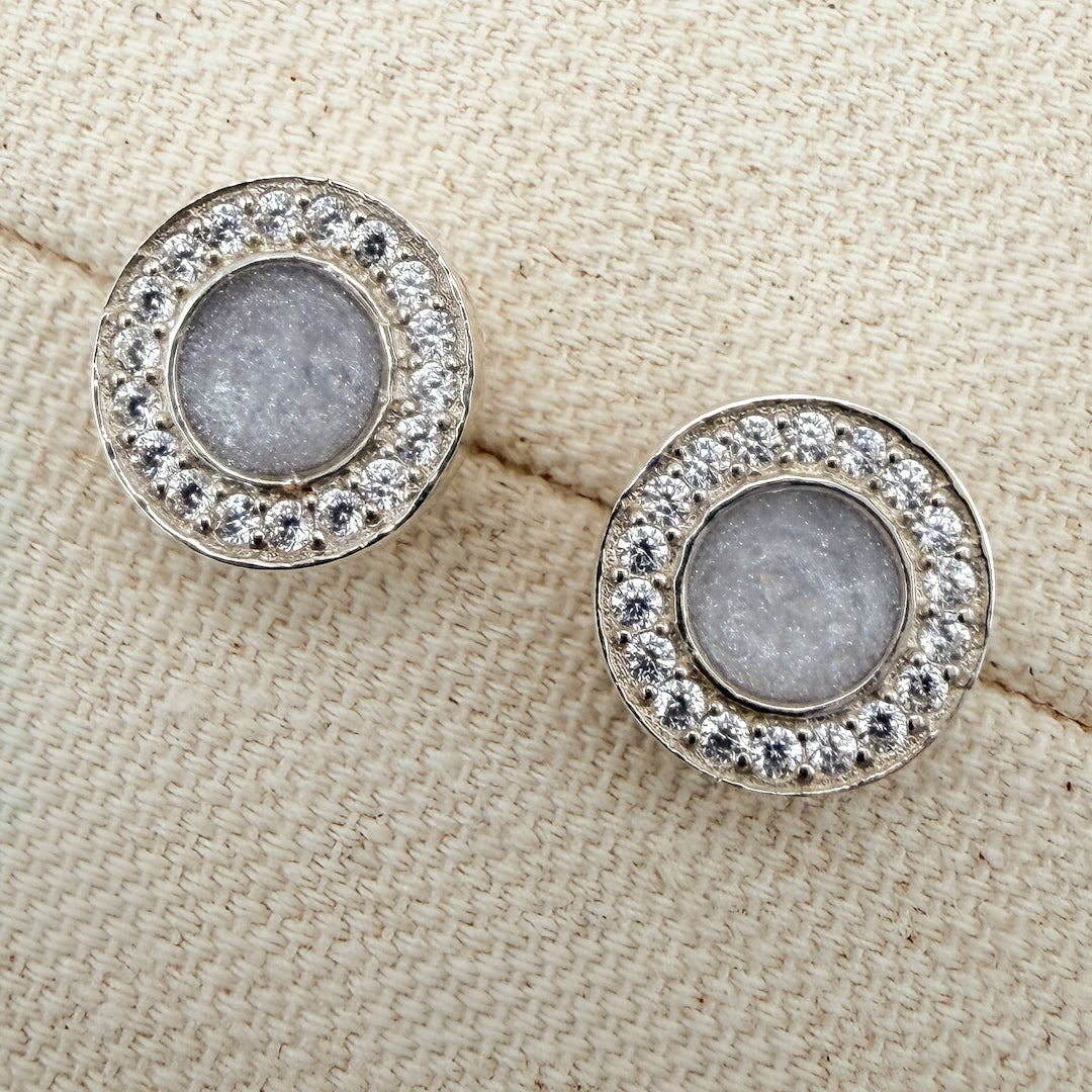A pair of round stud earrings surrounded by cubiic zirconia stones with a white silver centre featuring hidden hair inside. 