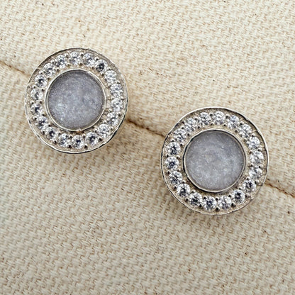 A pair of round stud earrings surrounded by cubiic zirconia stones with a white silver centre featuring hidden hair inside. 
