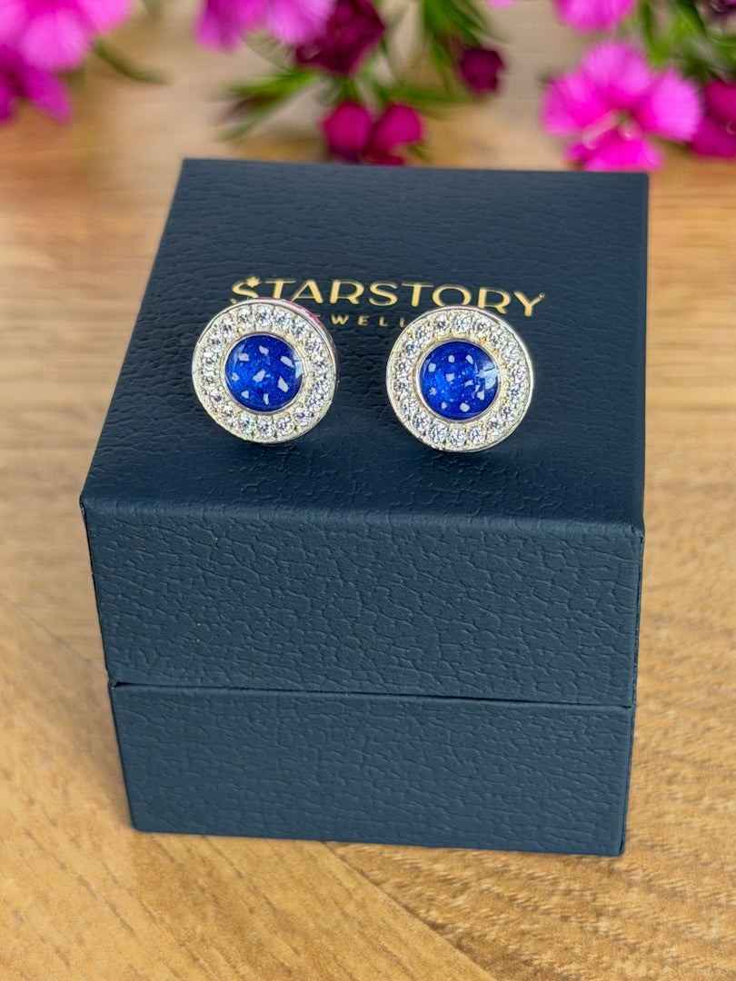 A pair of Starstory Jewellery stud earrings with round blue centres surrounded by clear cubic zirconia crystals in a silver-toned setting, displayed on a dark blue textured jewellry box on a wooden surface with blurred pink flowers in the background.