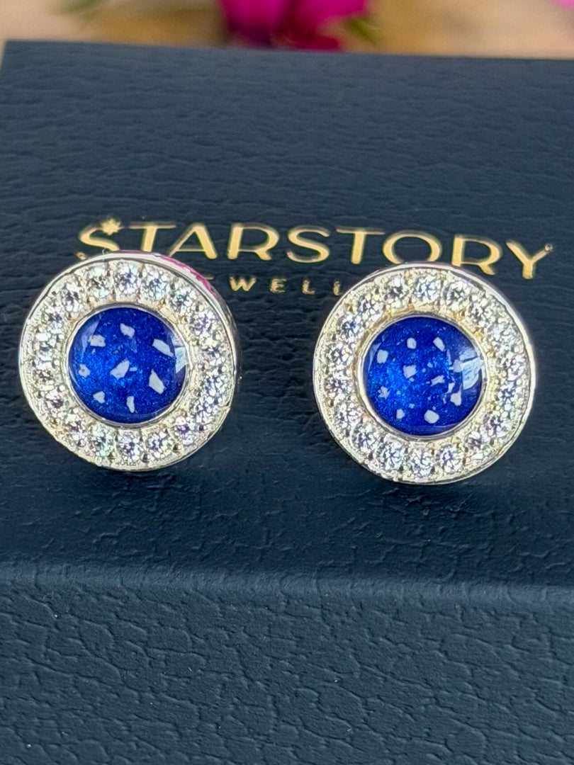 A pair of Starstory Jewellery stud earrings with round blue centres surrounded by clear cubic zirconia crystals in a silver-toned setting, displayed on a dark blue textured jewellry box on a wooden surface with blurred pink flowers in the background.