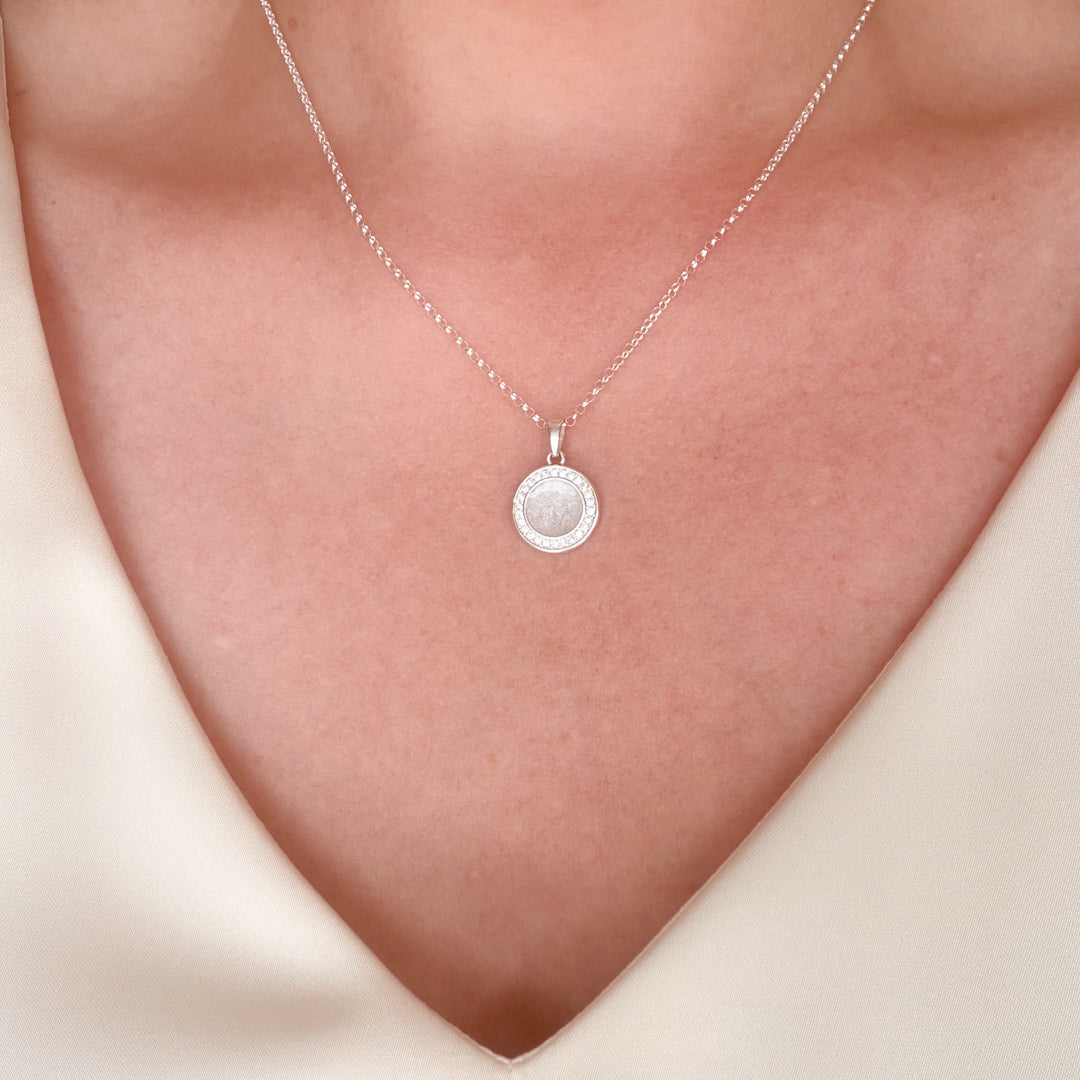 A close-up of a silver chain necklace with a round pendant worn on a person's neck. The pendant features a textured white silvery resin stone with hidden inclusion surrounded by a halo of small clear cubic zirconia stones, set against warm skin tones.