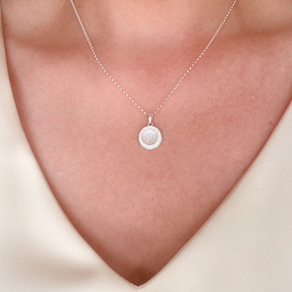 A close-up of a silver chain necklace with a round pendant worn on a person's neck. The pendant features a textured white silvery resin stone with hidden inclusion surrounded by a halo of small clear cubic zirconia stones, set against warm skin tones.
