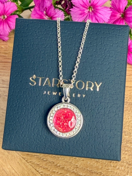 A round pendant necklace with a silver-toned setting, surrounded by cubic zirconia featuring a textured bright pink center stone with visible blended ashes, hanging from a silver chain against a dark textured background with gold 'Starstory Jewellery branding.