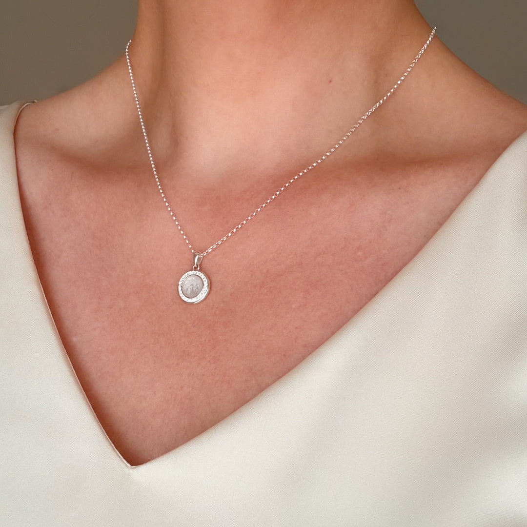 A close-up of a delicate silver chain necklace with a small circular pendant worn on a model's neck, paired with a cream-colored V-neck top.