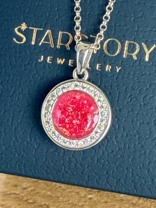 A round pendant necklace with a silver-toned setting, surrounded by cubic zirconia featuring a textured bright pink center stone with visible blended ashes, hanging from a silver chain against a dark textured background with gold 'Starstory Jewellery branding.