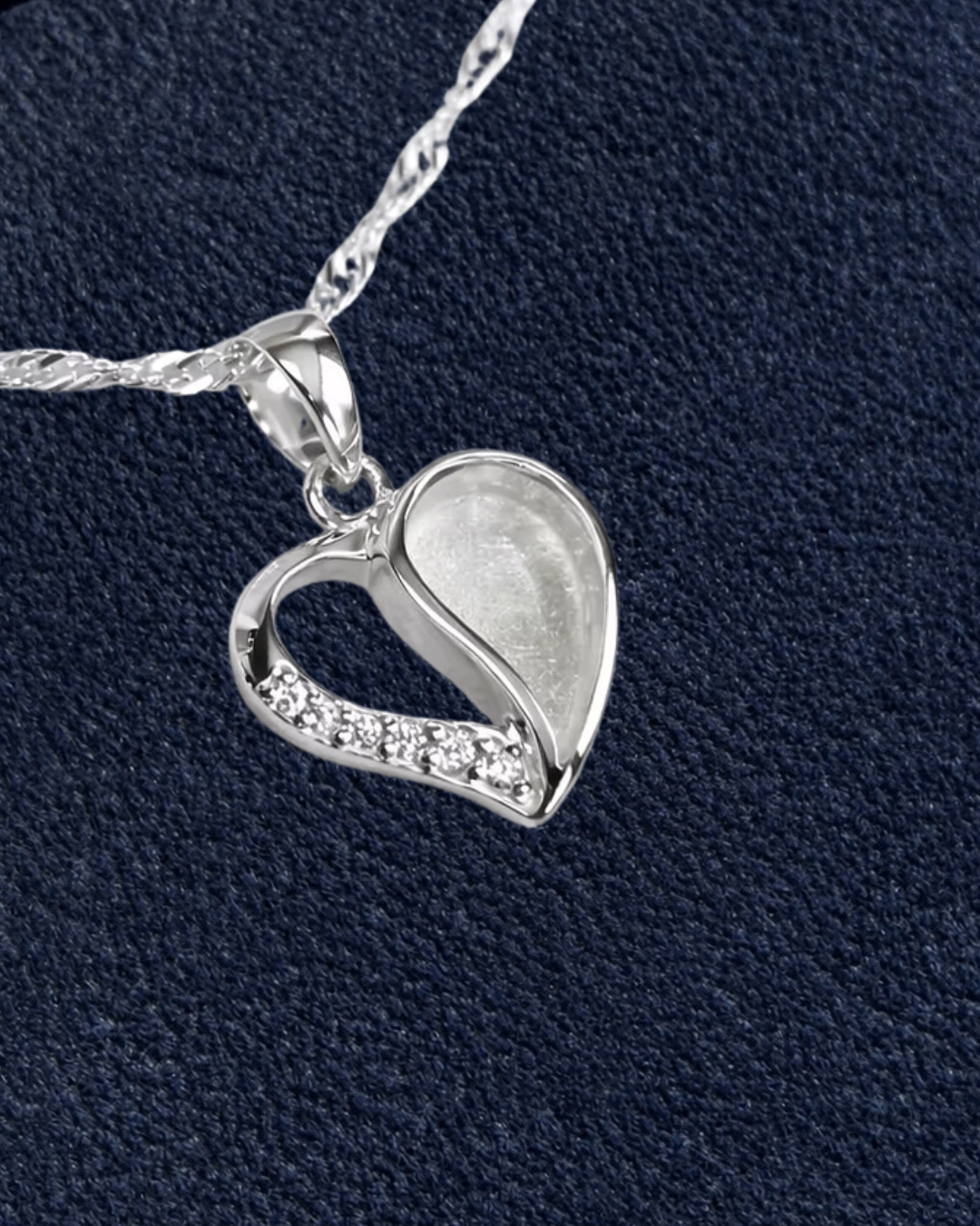 A sterling silver heart-shaped pendant necklace with a small row of clear cubic zirconia stones along one side, shown on a textured dark navy background with a cream circle in the top right containing the word 'New'.