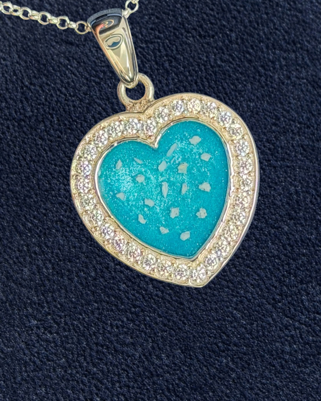 A heart-shaped pendant necklace featuring a bright turquoise blue center with tiny white flecks, surrounded by a gold border set with small clear stones, shown on a textured dark navy background.