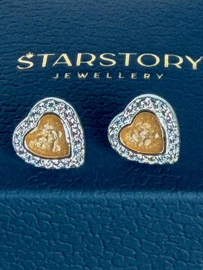 A pair of heart-shaped stud earrings surrounded by cubic zirconia and a yellow centre with visible ash. The earrings are displayed on a dark blue Starstory Jewellery box on a wooden surface with pink flowers in the background.