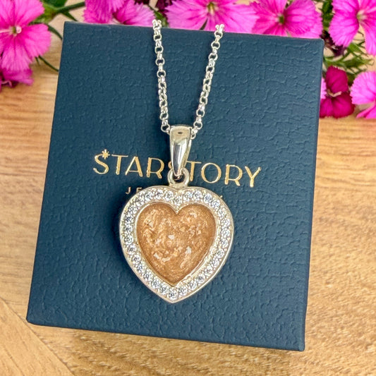 A heart-shaped pendant necklace with visible ashes inclusiondisplayed on a dark blue Starstory Jewellery box, set on a wooden surface with pink flowers in the background.
