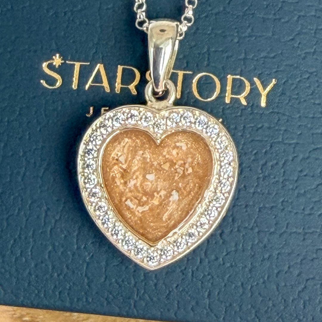 A heart-shaped pendant necklace with visible ashes inclusion displayed on a dark blue Starstory Jewellery box, set on a wooden surface with pink flowers in the background.