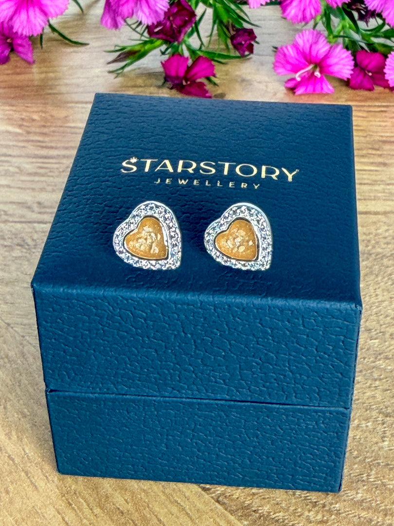 A pair of heart-shaped stud earrings surrounded by cubic zirconia and a yellow centre with visible ash. The earrings are displayed on a dark blue Starstory Jewellery box on a wooden surface with pink flowers in the background.