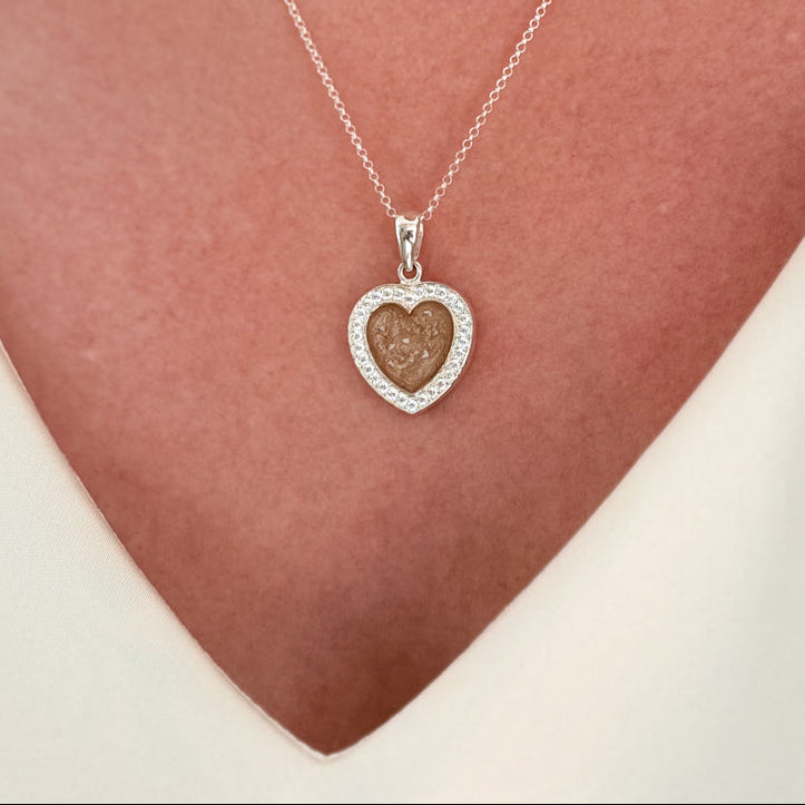 A close-up of a person wearing a delicate sterling silver chain necklace with a heart-shaped pendant with cubic zirconia stones and a champagne centre with visible ash. 