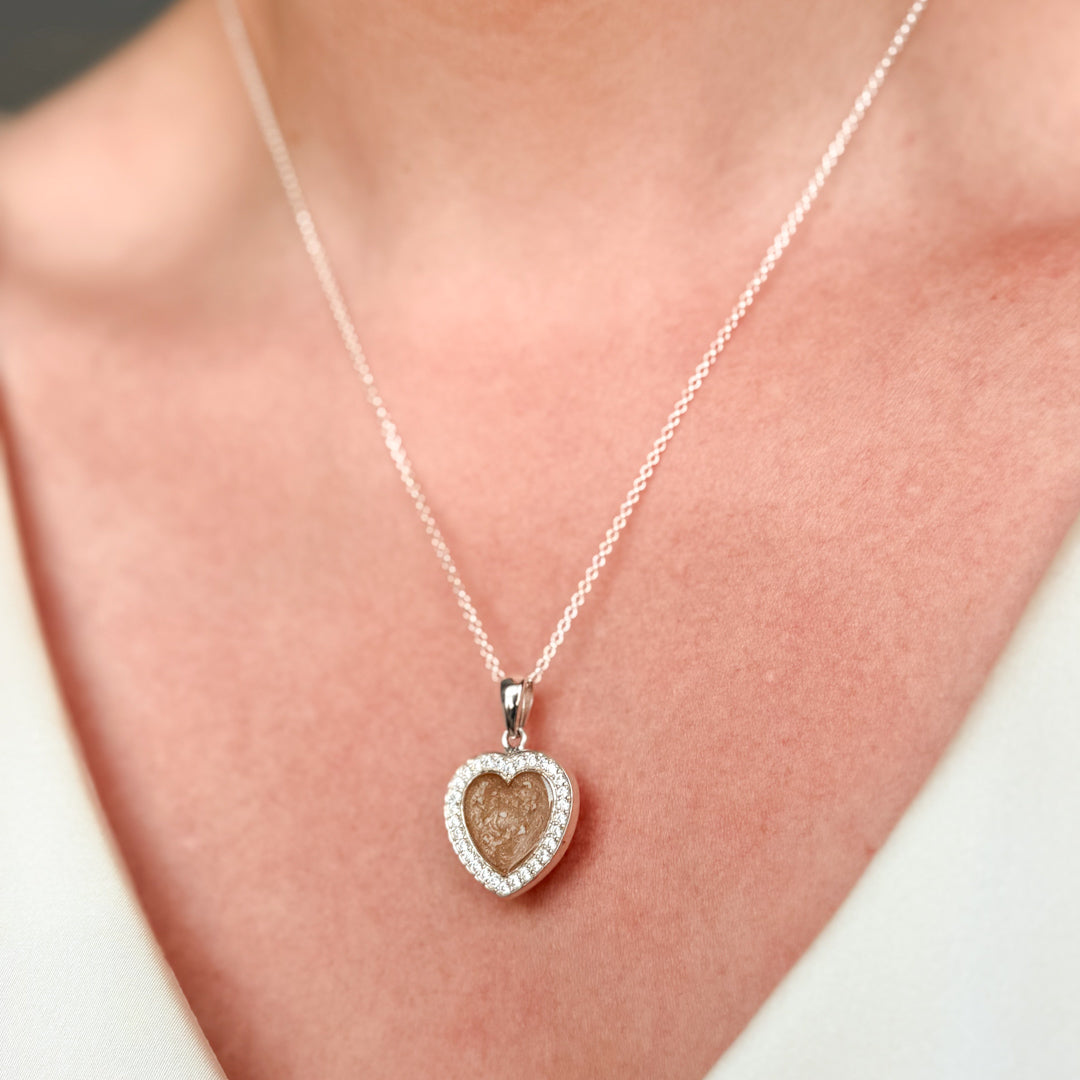 A close-up of a person wearing a delicate sterling silver chain necklace with a heart-shaped pendant with cubic zirconia stones and a champagne centre with visible ash. 