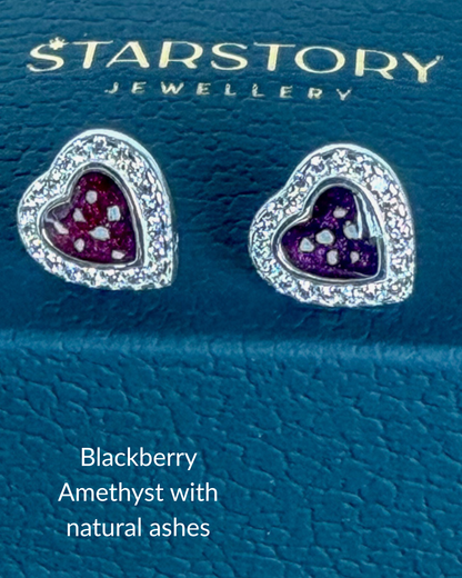 A pair of heart-shaped stud earrings by Starstory Jewellery, featuring a silver-tone halo border with clear cubic zirconia stones and a central purple amethyst heart with small ashes inclusions. 